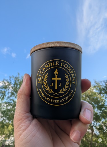 Black candle container with gold logo and text held against a blue sky with trees.