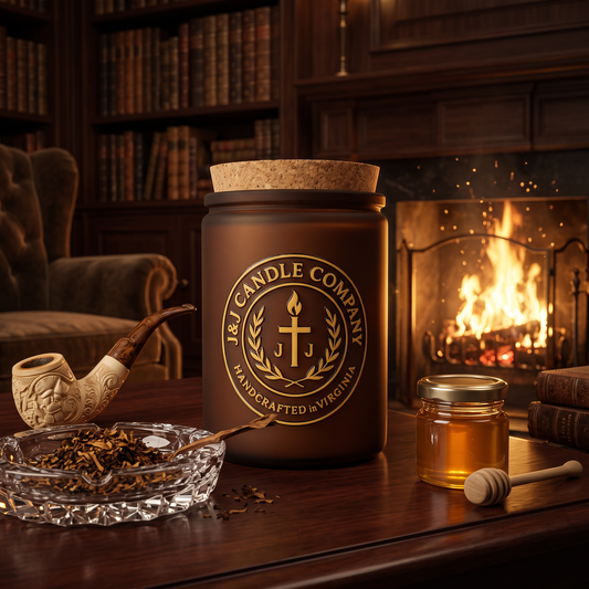 A large amber-brown jar candle with a cork lid and a gold circular logo reading 'J&L Candle Company' stands on a wooden table in a cozy room with a burning fireplace, books, a pipe, and a small honey jar nearby.