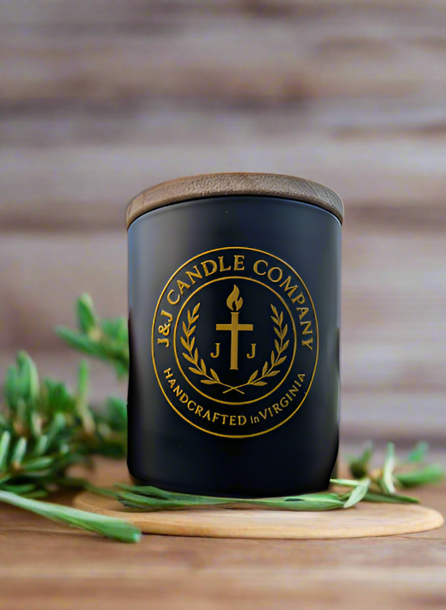Black candle jar with wooden lid and J&J Candle Company logo on a wooden surface with greenery.